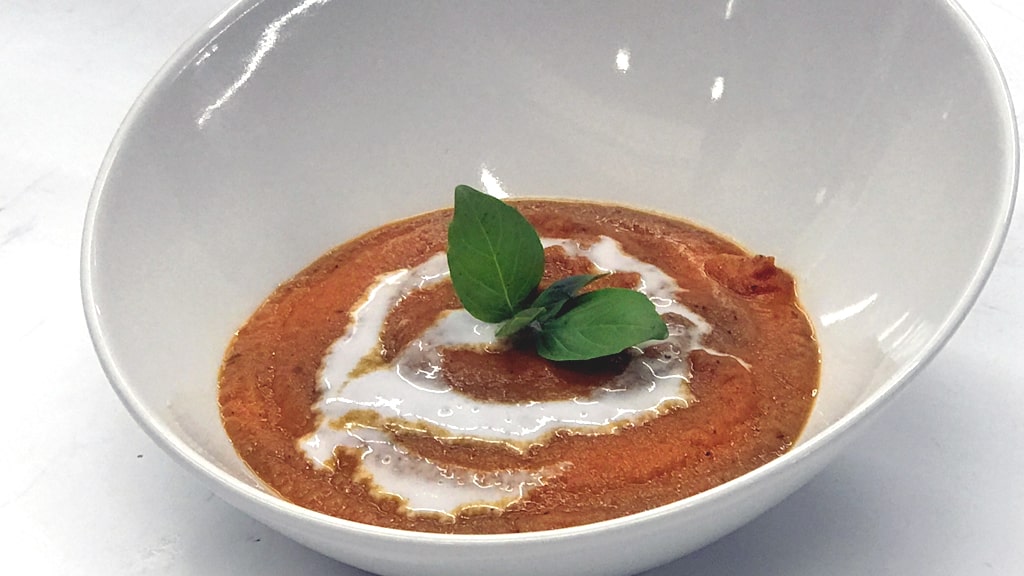The finished bowl of tomato soup with a coconut cream drizzle and a basil garnish.