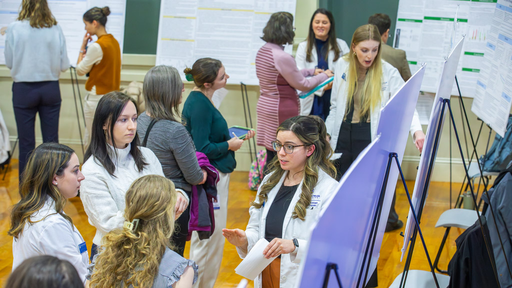 several white-coated PA students explain their poster projects while Scholarship Day guests listen