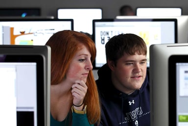 Two JWU students in front of a bank of computer display screens.