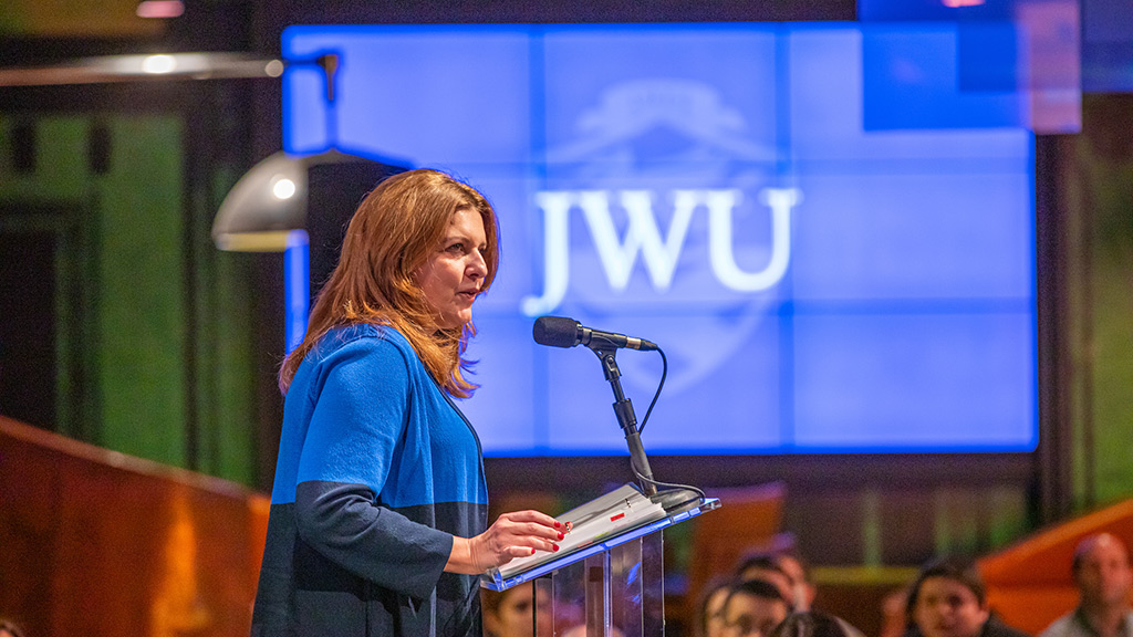JWU Campus President Marie Bernardo Sousa speaking at a podium with a JWU logo in the background