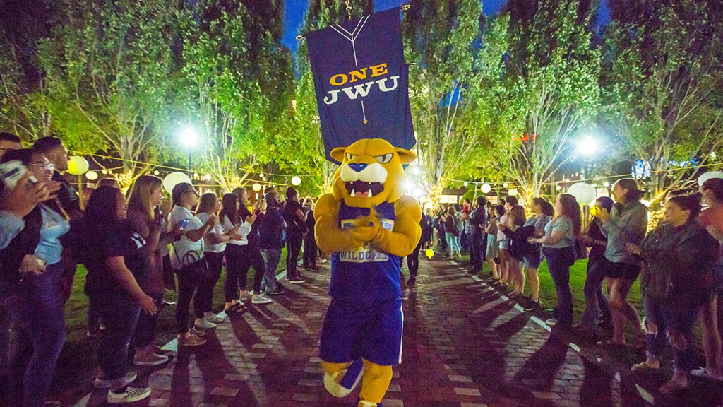 Wildcat Willie surrounded by students at Ignite the Night with a banner behind him that says One JWU