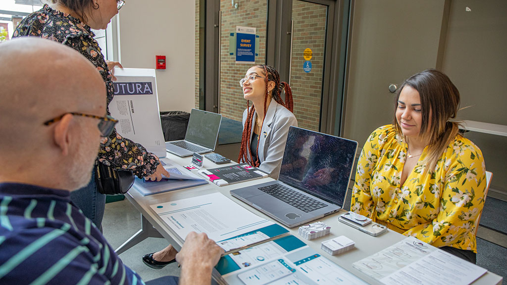 Students sitting with their portfolio books while members of the JWU community look at the designs