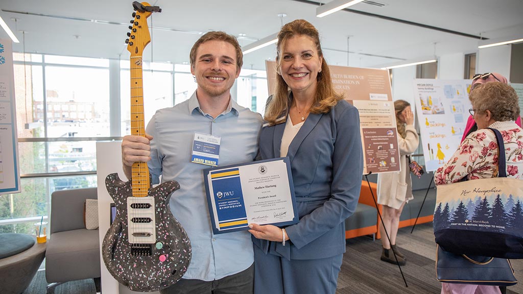 Matthew Hartung ’24 poses with his Infinity Guitar while being presented a symposium award by JWU Providence Campus President Marie Bernardo-Sousa