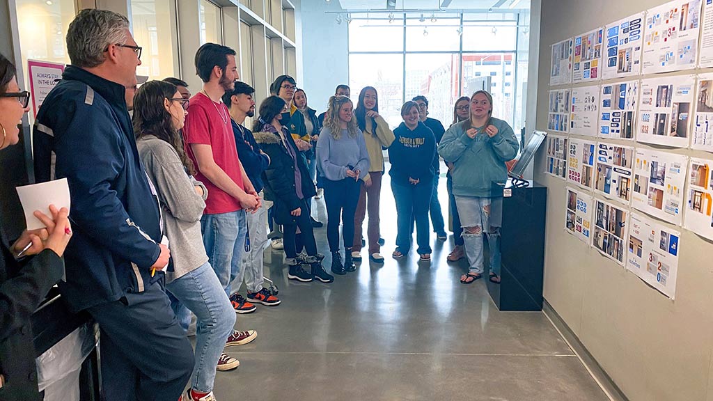 Wayfinding class design winner Shea Lambert, far right, talks about her design to a group of people in an exhibit hallway