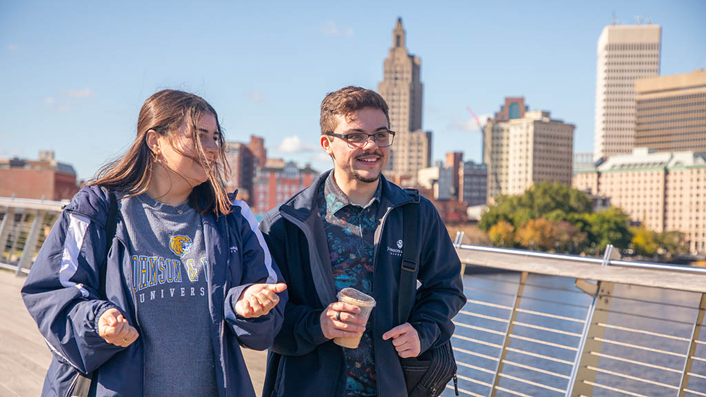 a photo of Lucia Manzo '25 and Jae Grenier '23 walking outdoors on the pedestrian bridge against a Providence skyline