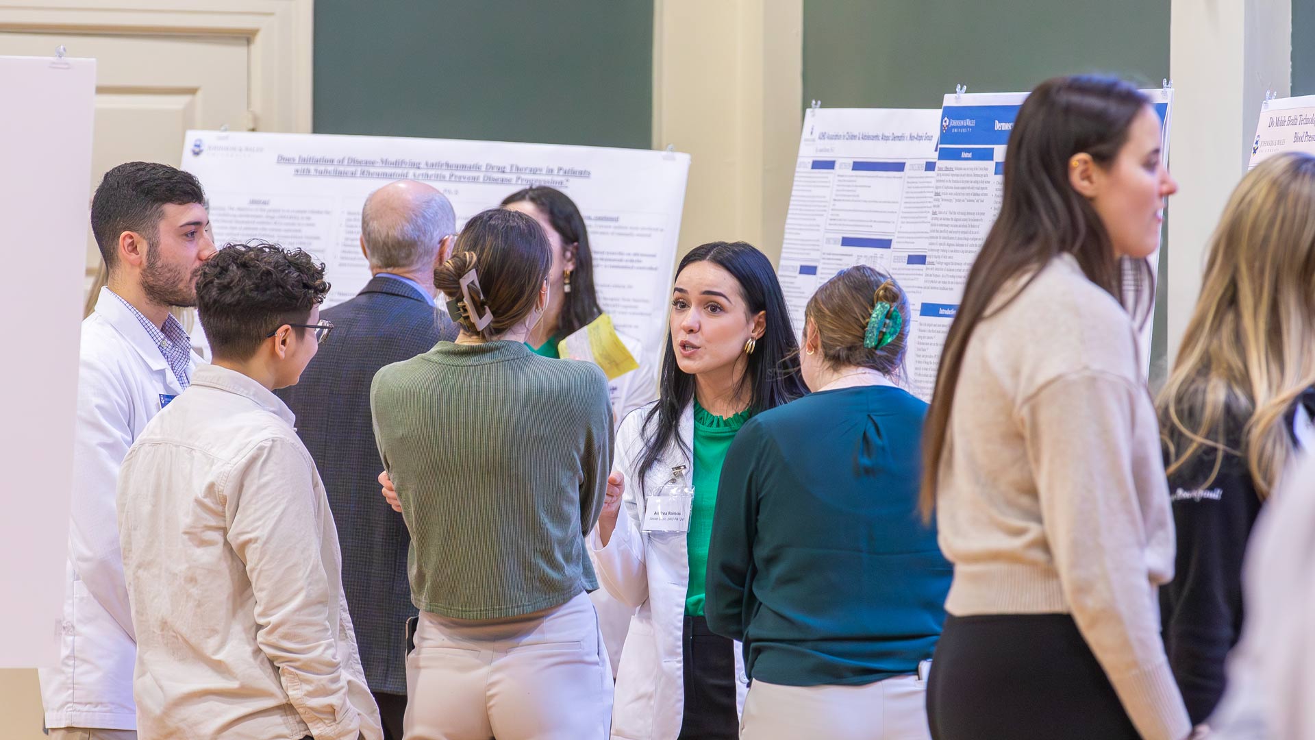 JWU PA students in white coats take turns explaining their master's research to guests at 2024 Scholarship Day