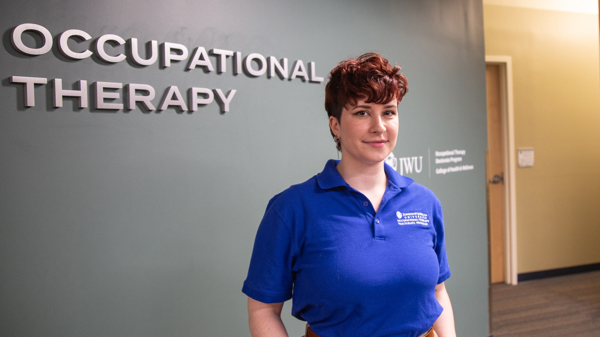 a photo of Molly Gould OTD '24 standing in front of the sign for the Occupational Therapy program space at JWU