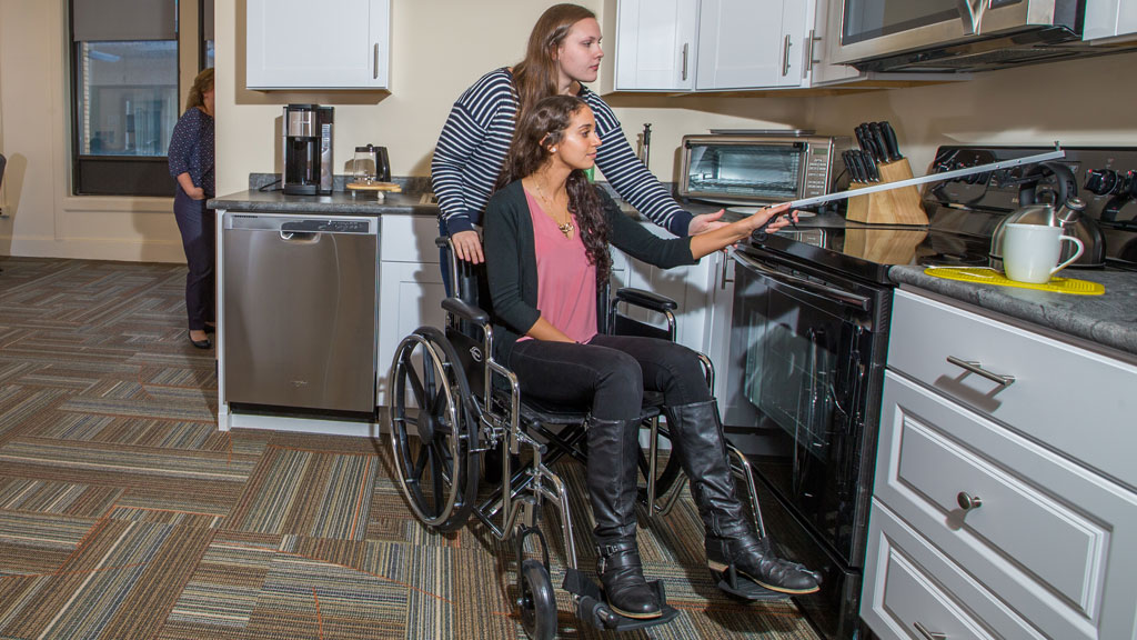 two female JWU students test out the OTD lab space, one posing in a wheelchair with an adaptive device in hand