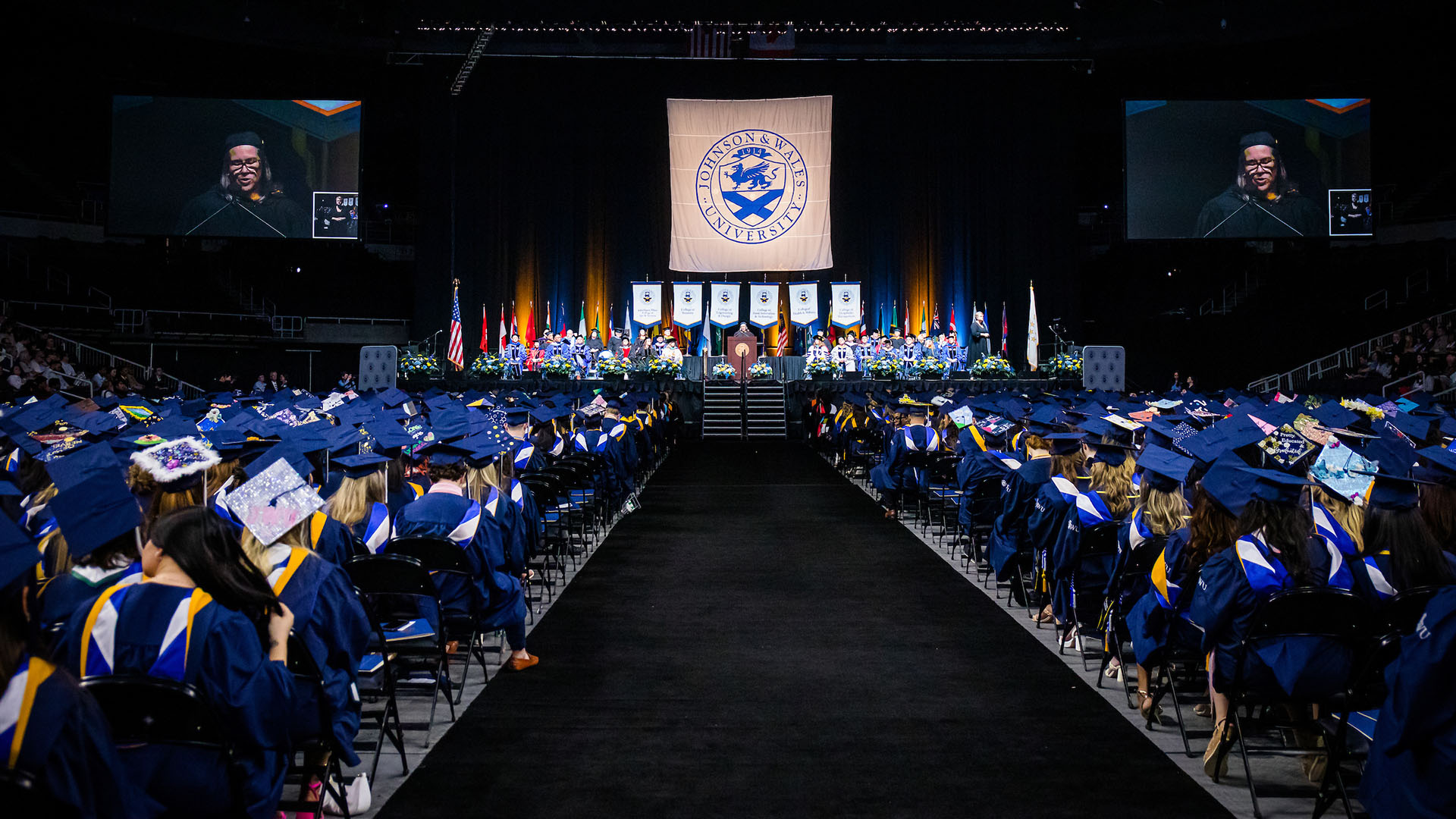 An audience of college graduate looking up at a person speaking on a stage.