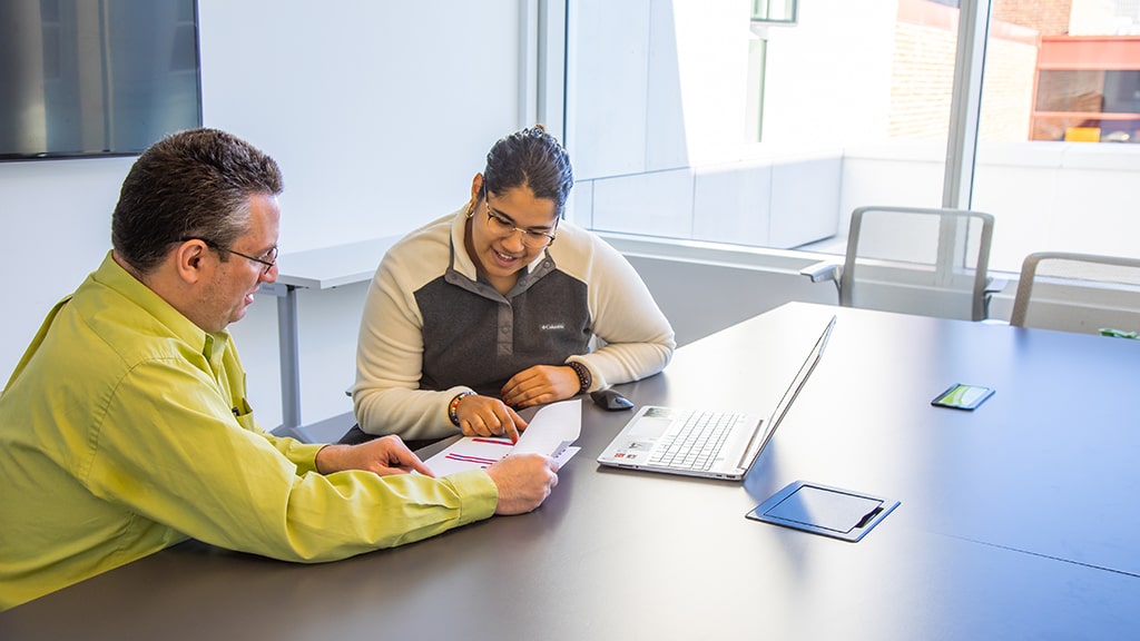 An academic advisor sits with a student at a desk and shows them information on paper
