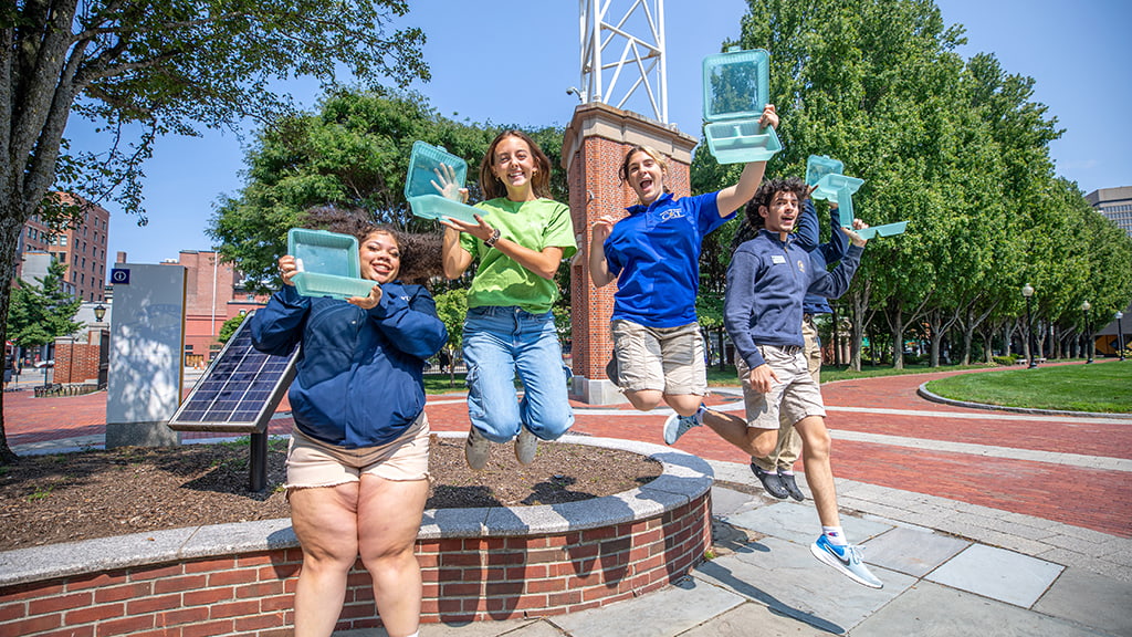 Students jump off brick wall holding JWU's reusable takeout containers