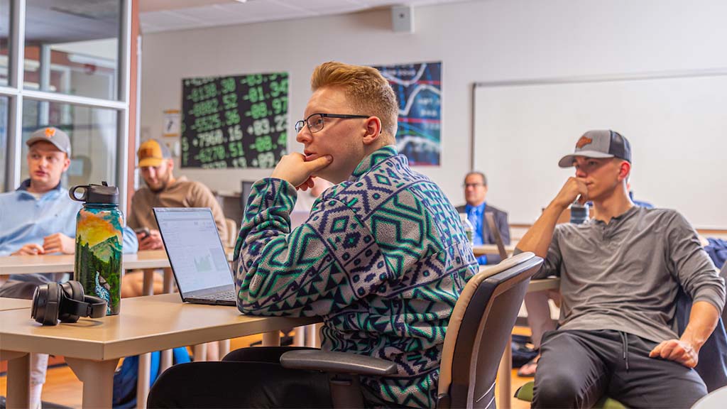 JWU Providence students seated in a classroom, listening attentively during a lesson. One student in the foreground works on a laptop while others sit behind him.