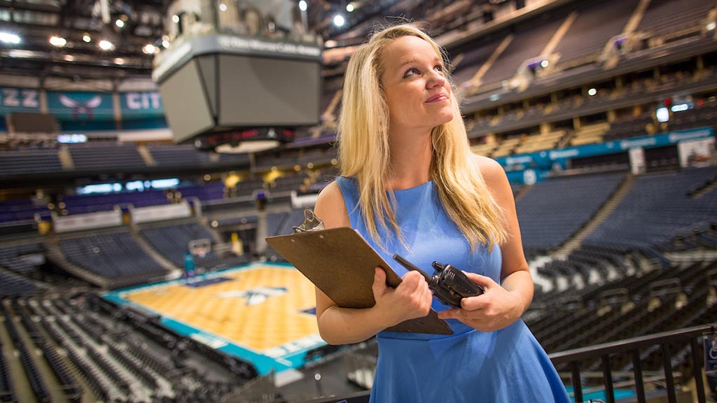 Sports management professional shown in a major arena venue with clipboard and walkie-talkie.