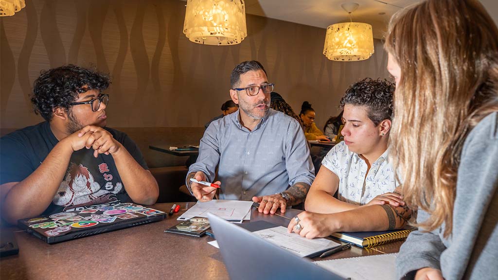 A JWU faculty member sits with three students at a table, guiding them through notes and assignments during a collaborative study session.