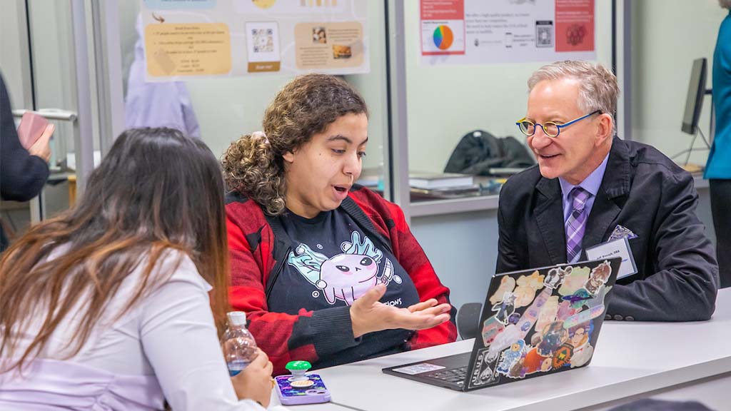 A JWU student speaks while showing something on a laptop to two others, including a faculty member, during a classroom discussion.