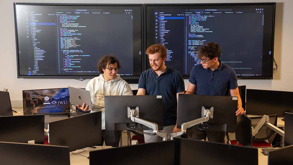 Three students collaborating in a high-tech workspace with multiple computer monitors displaying code.