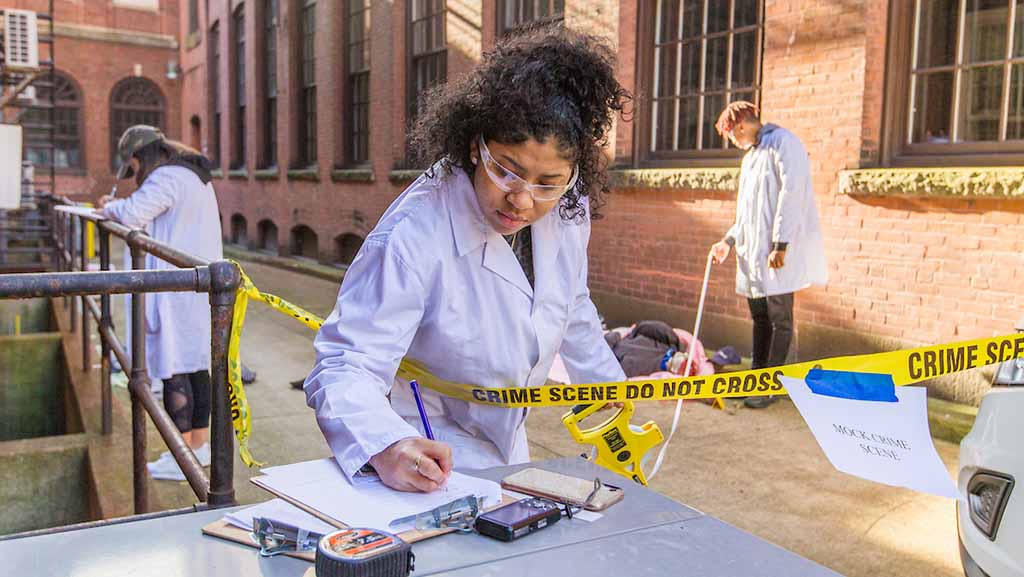 Person in a white lab coat working at a crime scene investigation, taking notes near "CRIME SCENE DO NOT CROSS" tape outside a brick building. Various forensic tools are visible on the table.