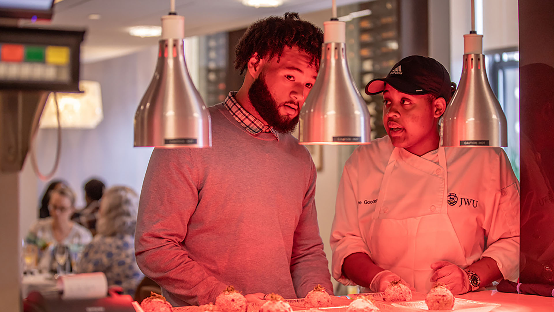 Two students standing near a heat lamp in a kitchen.