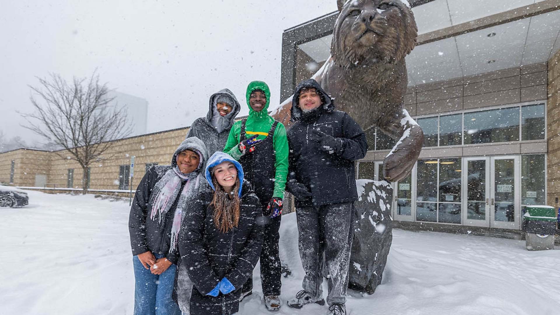 A group of people smiling and posing in the snow in front of the Wildcat statue.
