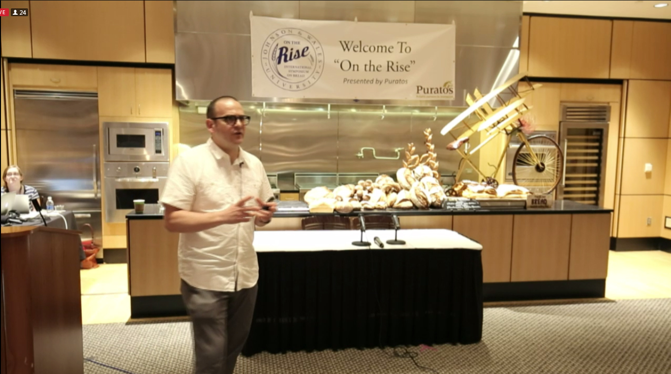 A man speaking in the JWU Amphitheater with bread art behind him