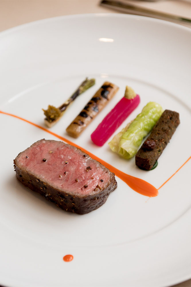 A seared meat medallion carefully plated with colorful vegetables and an orange sauce drizzle on a white plate.
