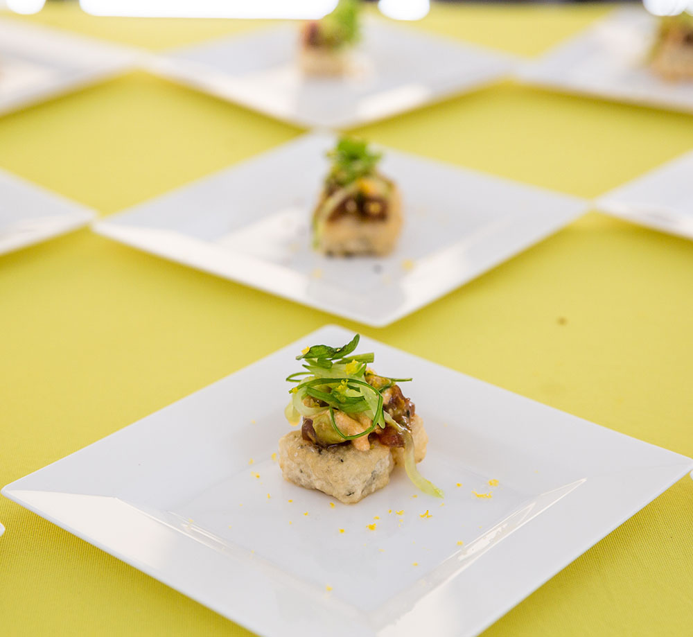 Artfully plated food on white square plates atop a bright yellow tablecloth.