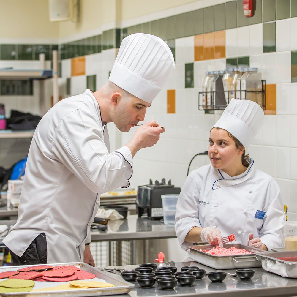 A professor tastes a student's dish in a culinary lab.