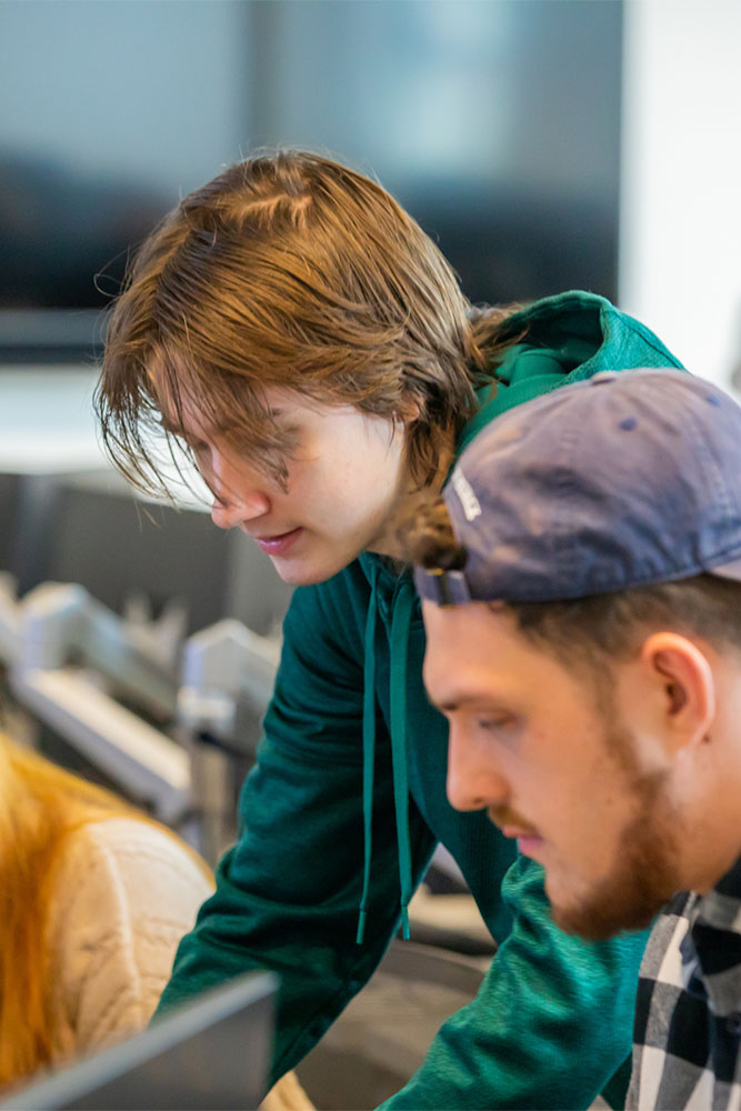 Two students collaborate in a computer lab, with one student leaning over to assist the other who is focused on a screen.