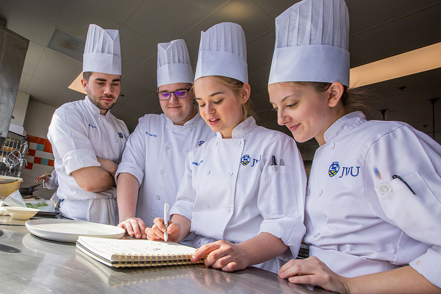 Four individuals in chef uniforms gather around a kitchen counter, examining a notebook together.