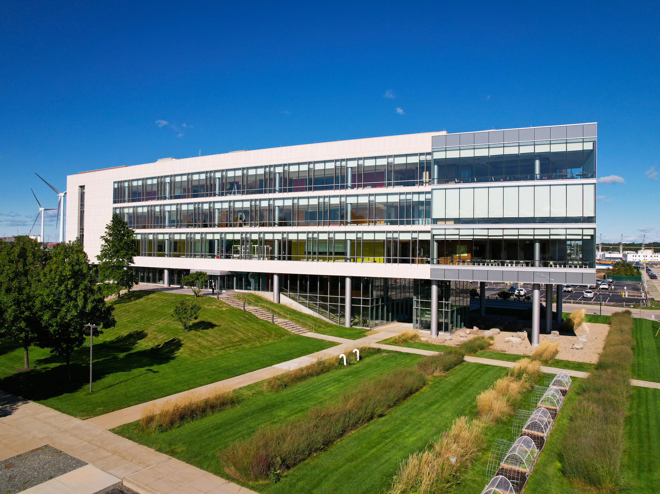 A modern multi-story academic building with large glass windows overlooks landscaped green lawns and walking paths, with wind turbines visible in the background under a clear blue sky.