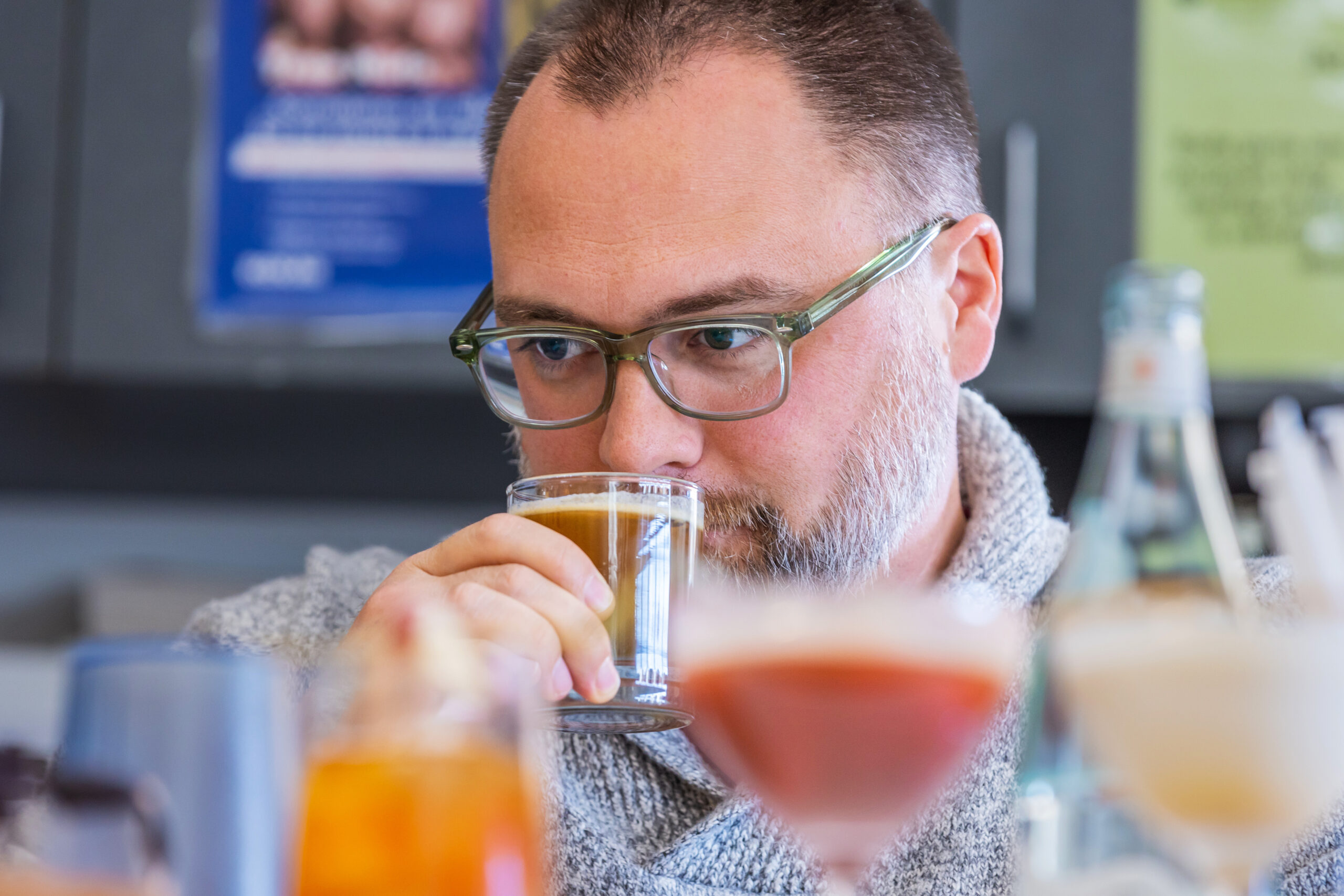A person wearing glasses closely examines and smells a small glass of beer, with other tasting glasses blurred in the foreground.