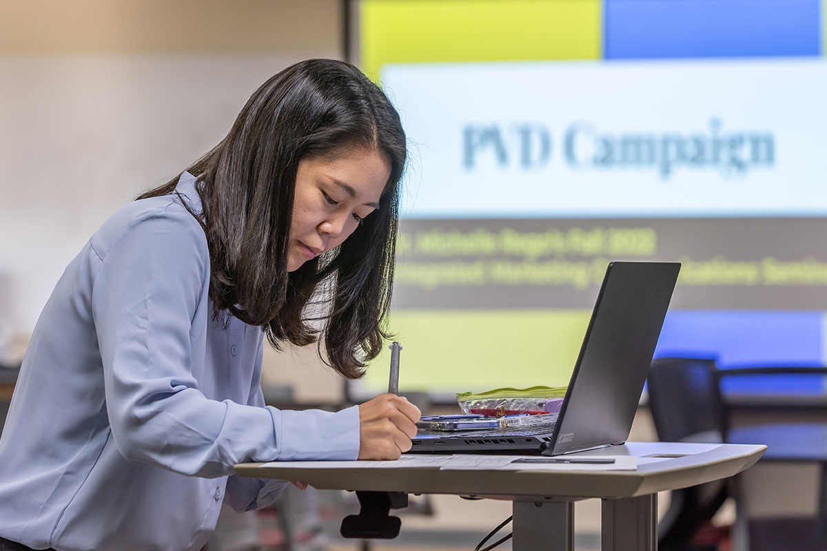 A student writing notes while seated at a desk with a laptop, in front of a presentation screen displaying "PVD Campaign."
