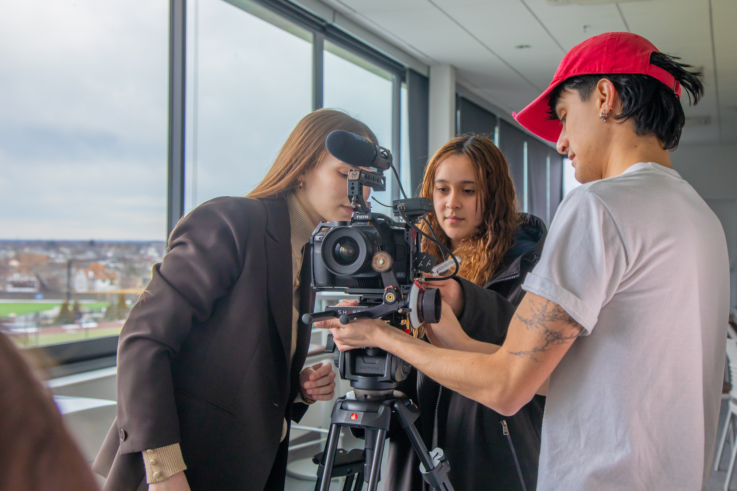 Three individuals collaborate closely around a professional camera mounted on a tripod in a brightly lit indoor space with large windows.