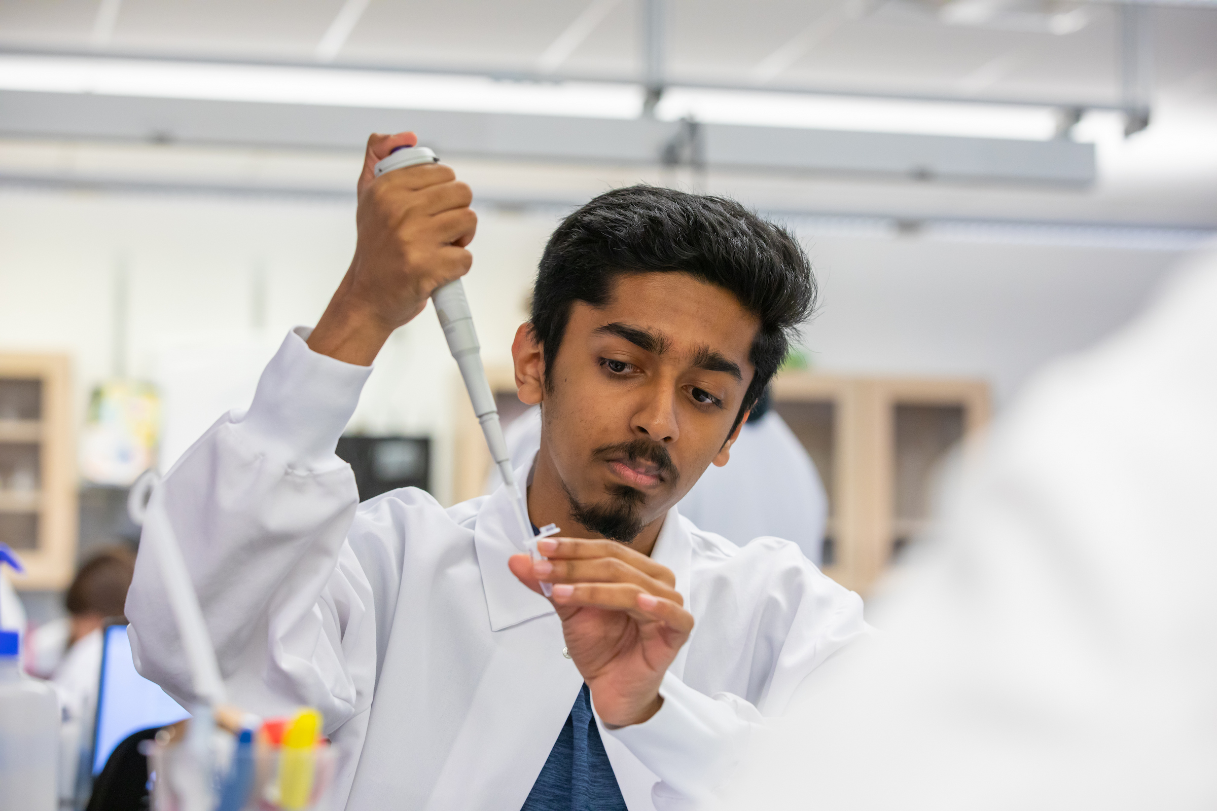 A student in a lab coat carefully uses a pipette to transfer liquid into a small test tube.