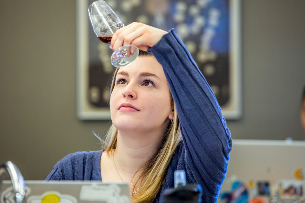 A person with blonde hair, wearing a blue sweater, holds a wine glass up to examine its contents while looking upward.