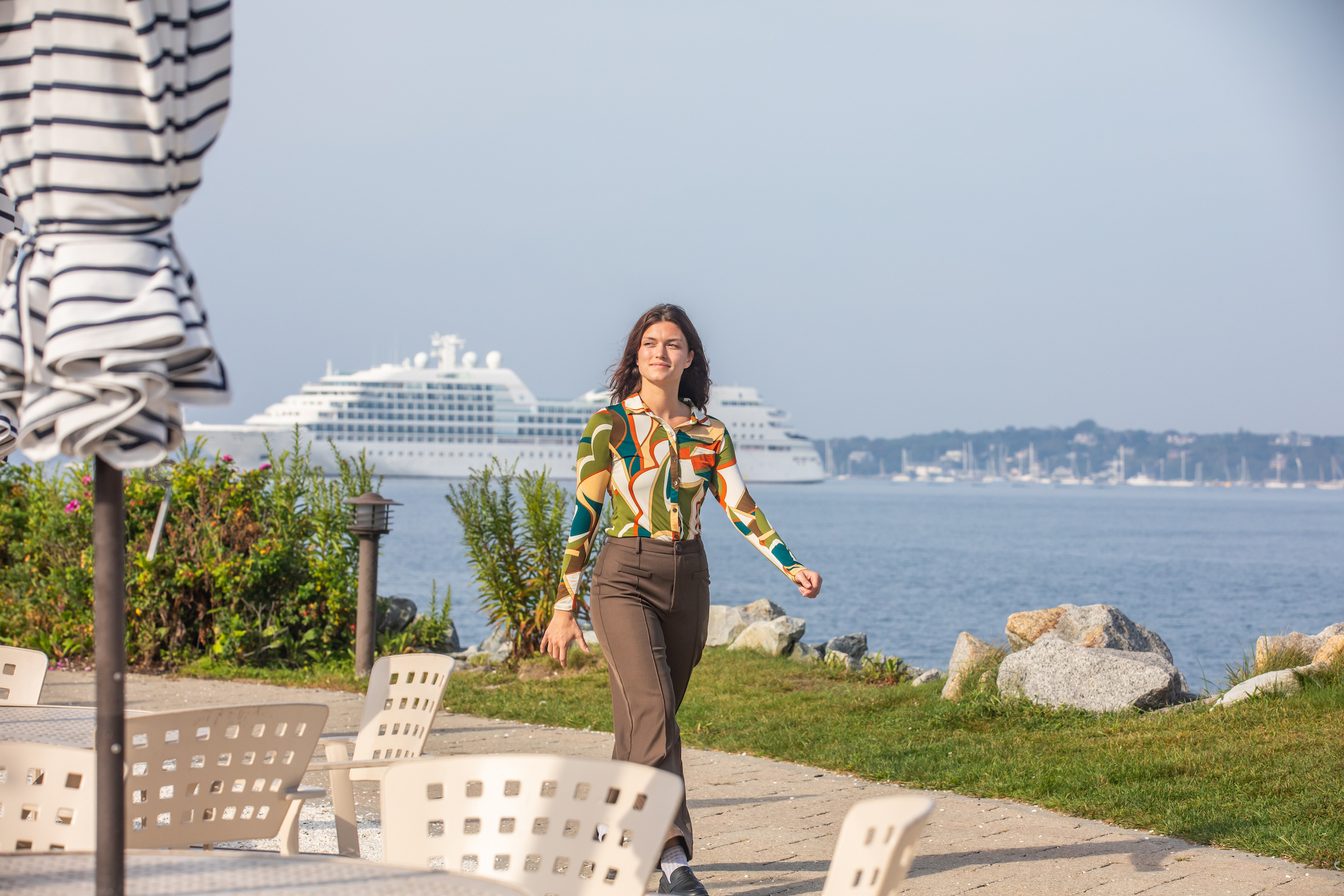 A person in patterned top and brown pants walks along a waterfront path with a cruise ship in the background.