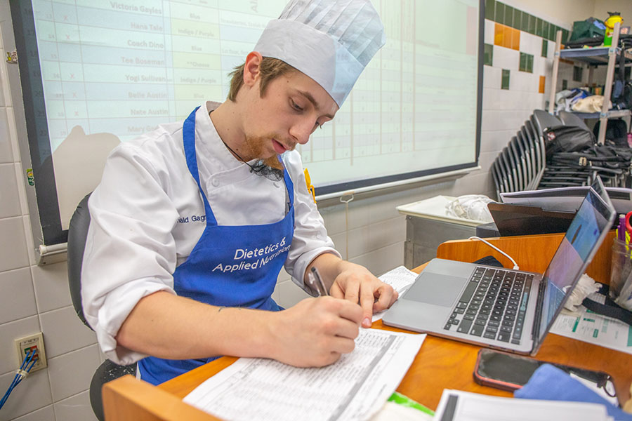 A student in a chef's uniform and blue apron writes at a desk with a laptop nearby.
