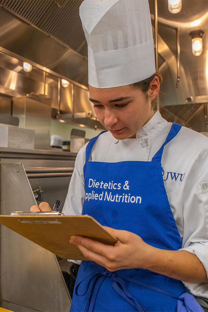A student in a chef's uniform and blue apron holds a clipboard in a kitchen.