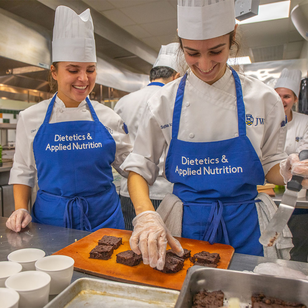 Two students in chef uniforms and aprons smile as they prepare brownies in a kitchen.