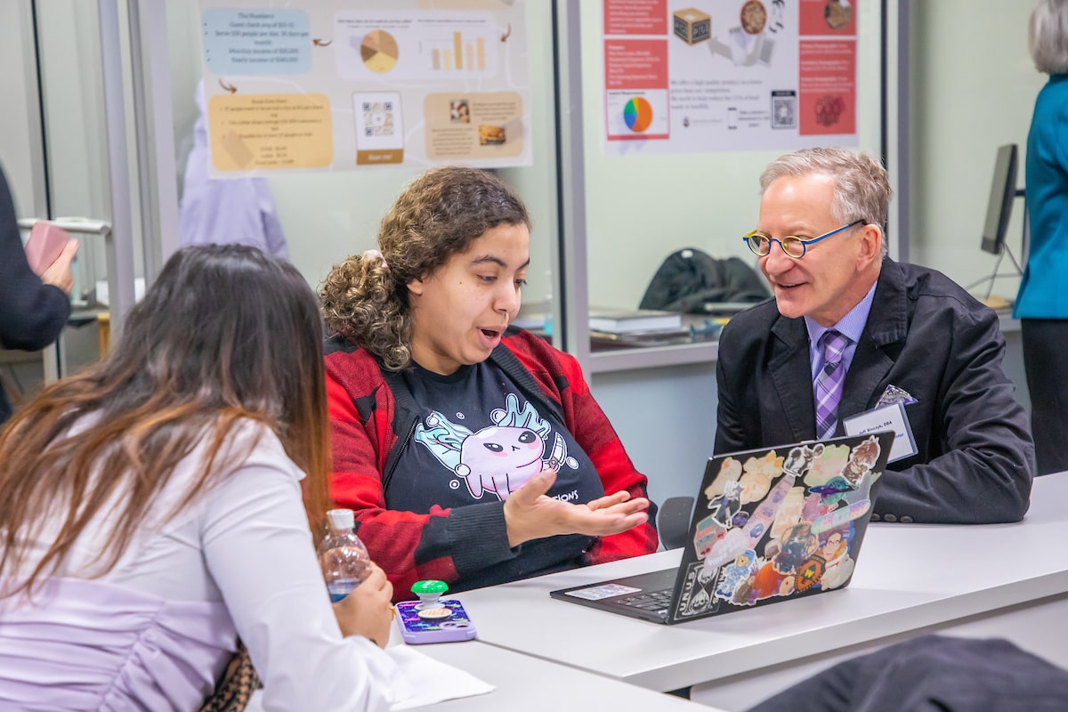 Two students sitting with a professor at a desk, looking at a laptop in a classroom.