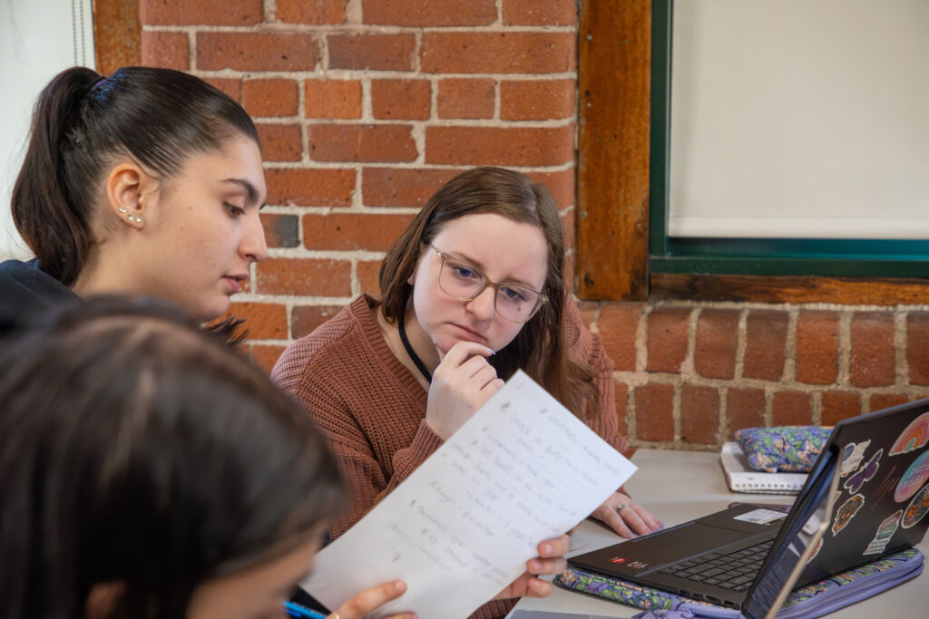 Two individuals collaborate closely over handwritten notes at a table, with a laptop and drinks nearby, set against a brick wall backdrop.
