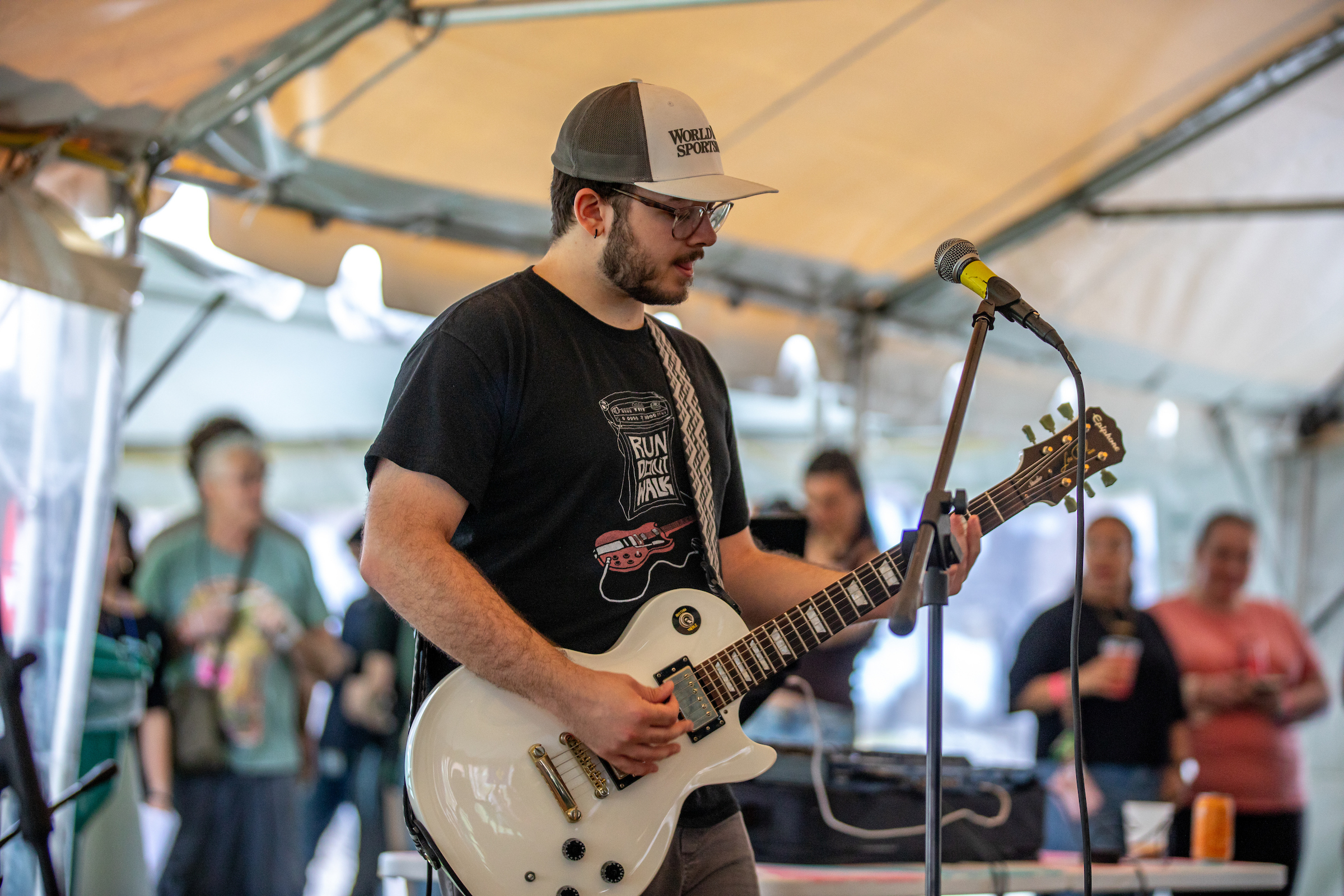 Annual SEEM Concert event a person plays a white electric guitar on stage under a tent, wearing a cap and glasses, with an audience in the background.
