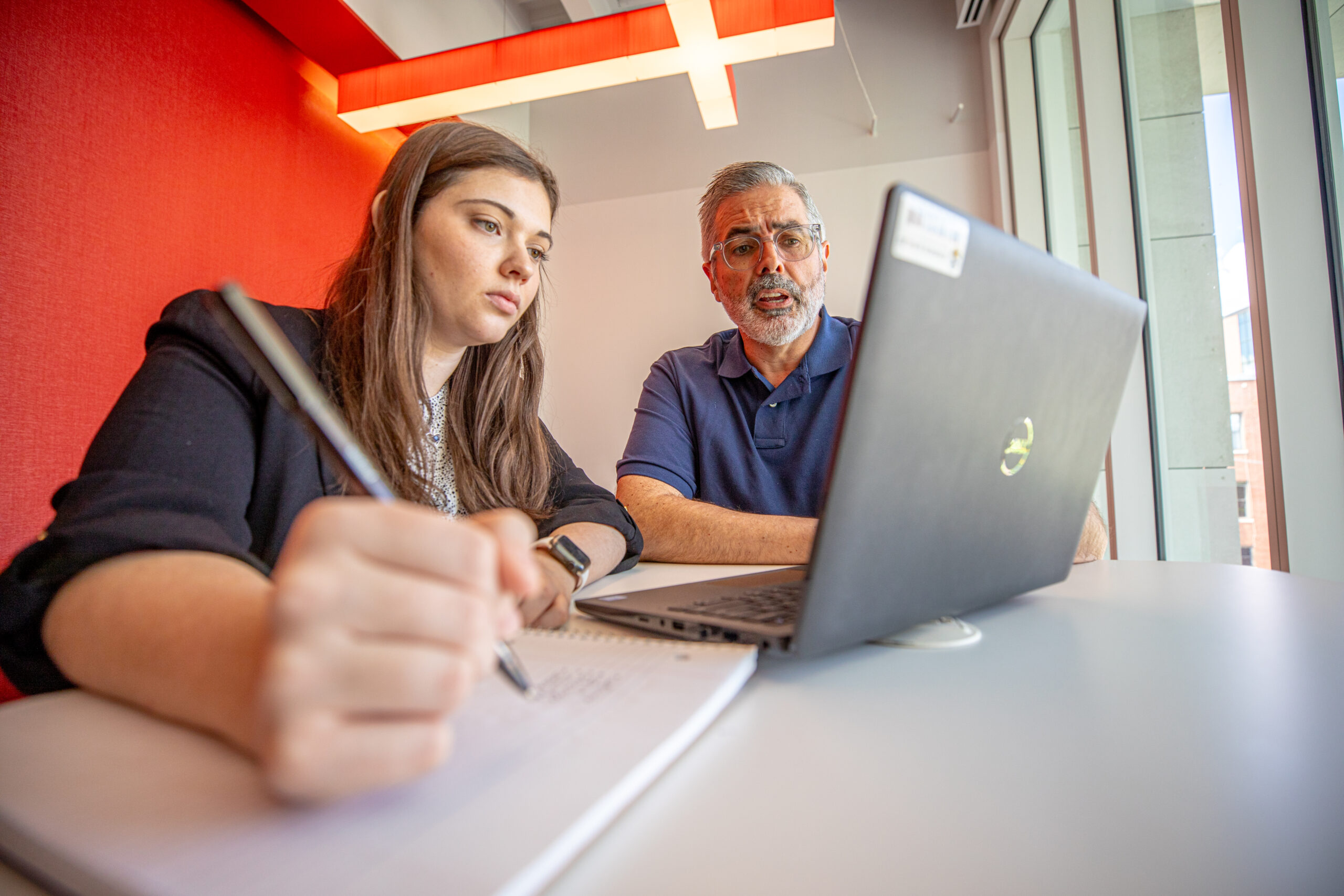 A student writes in a notebook while an advisor sits beside them, both focused on a laptop screen.