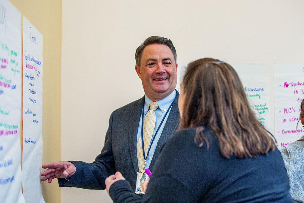 A person in a suit gestures toward a whiteboard while engaging in a discussion with a student in a classroom.
