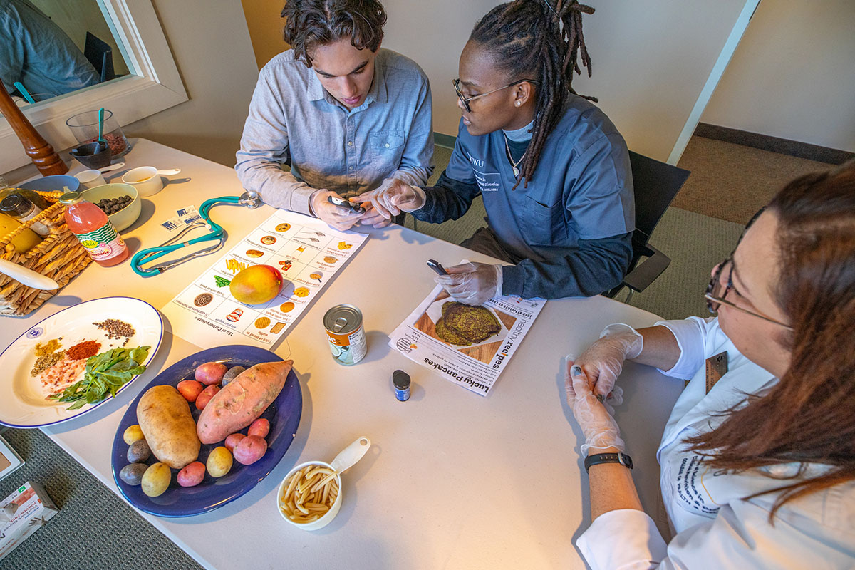 Three students sit at a table filled with food items and educational materials, examining nutrition content together.