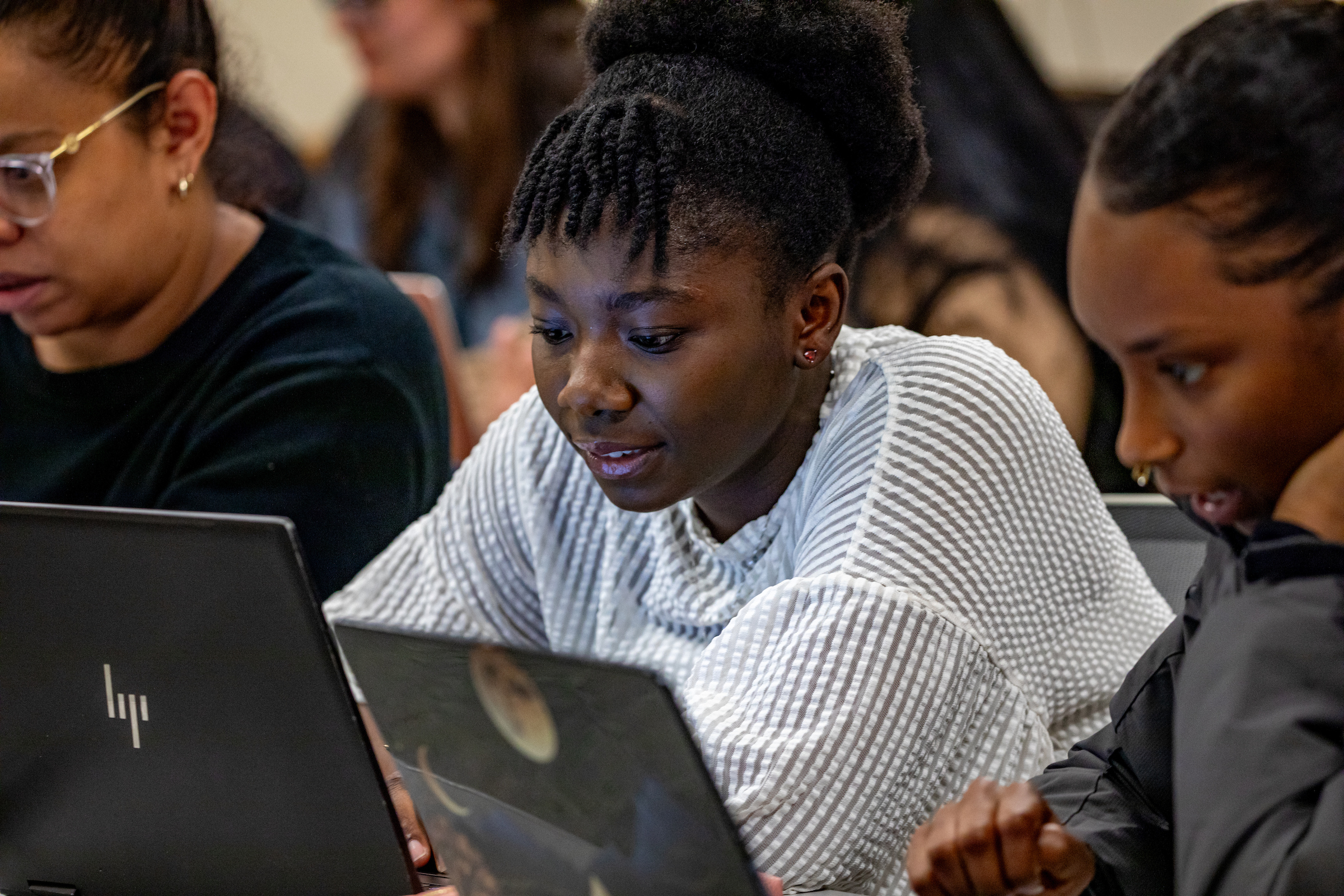 Three individuals are seated at laptops in a classroom setting, focused on their screens.