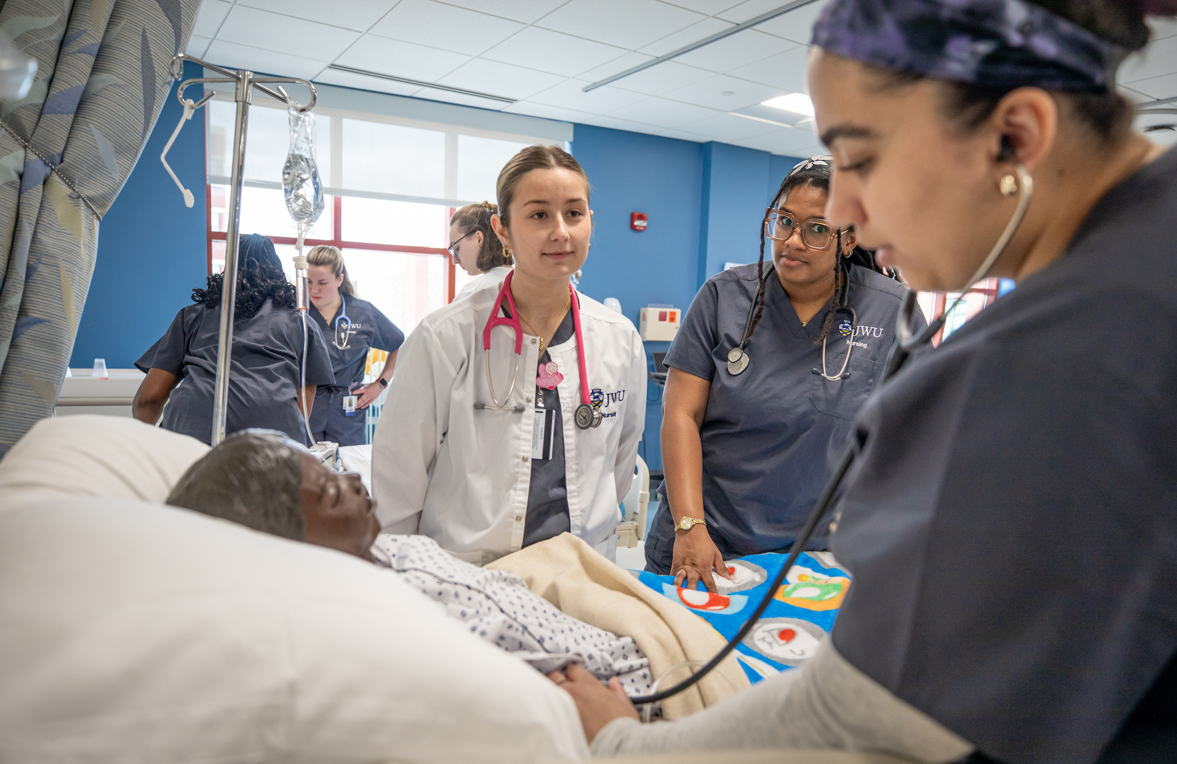 Medical trainees in a simulation lab attend to a patient in a hospital bed while others observe.