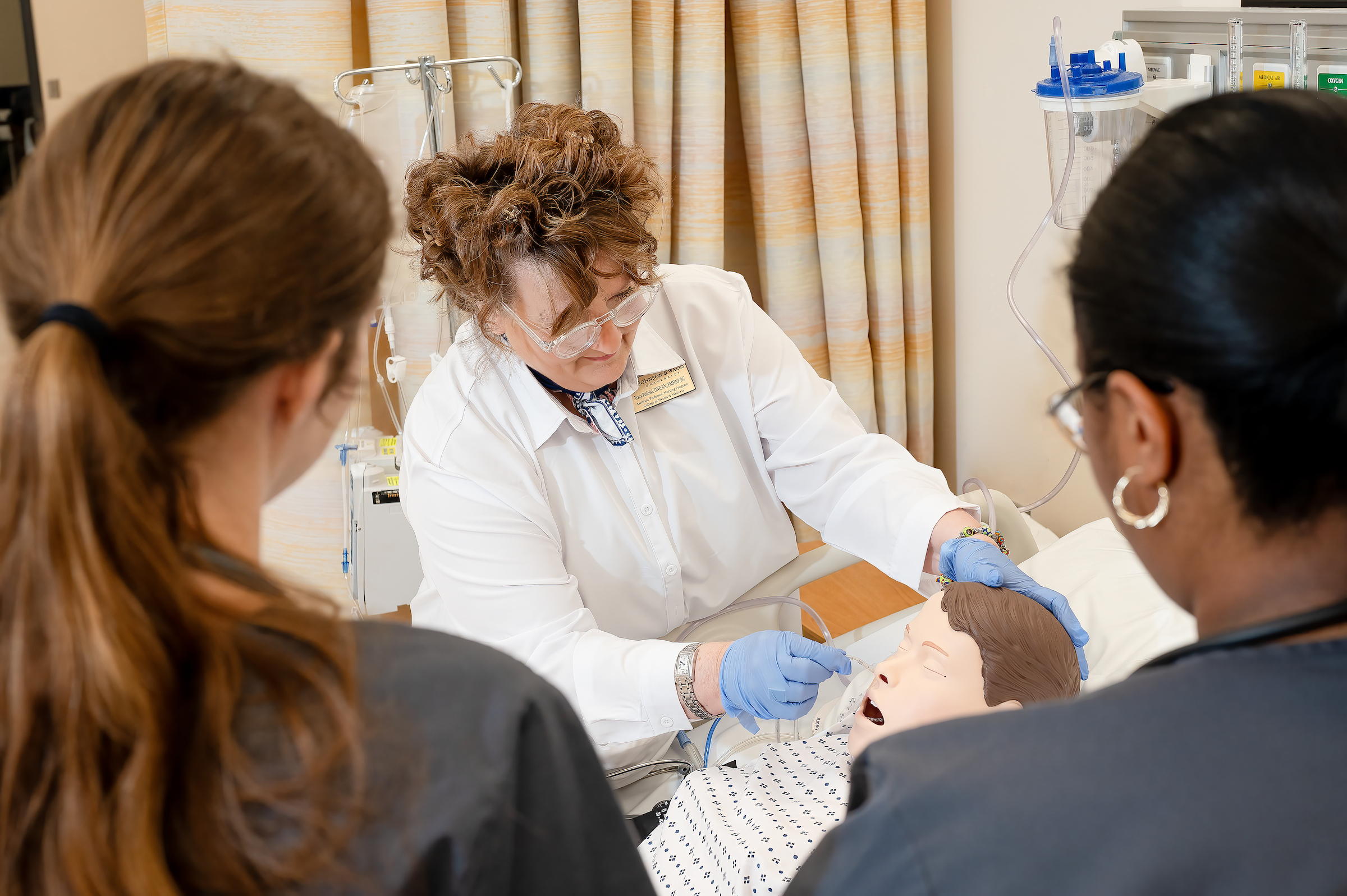 A healthcare instructor demonstrates a procedure on a medical mannequin while two students observe closely in a clinical training setting.