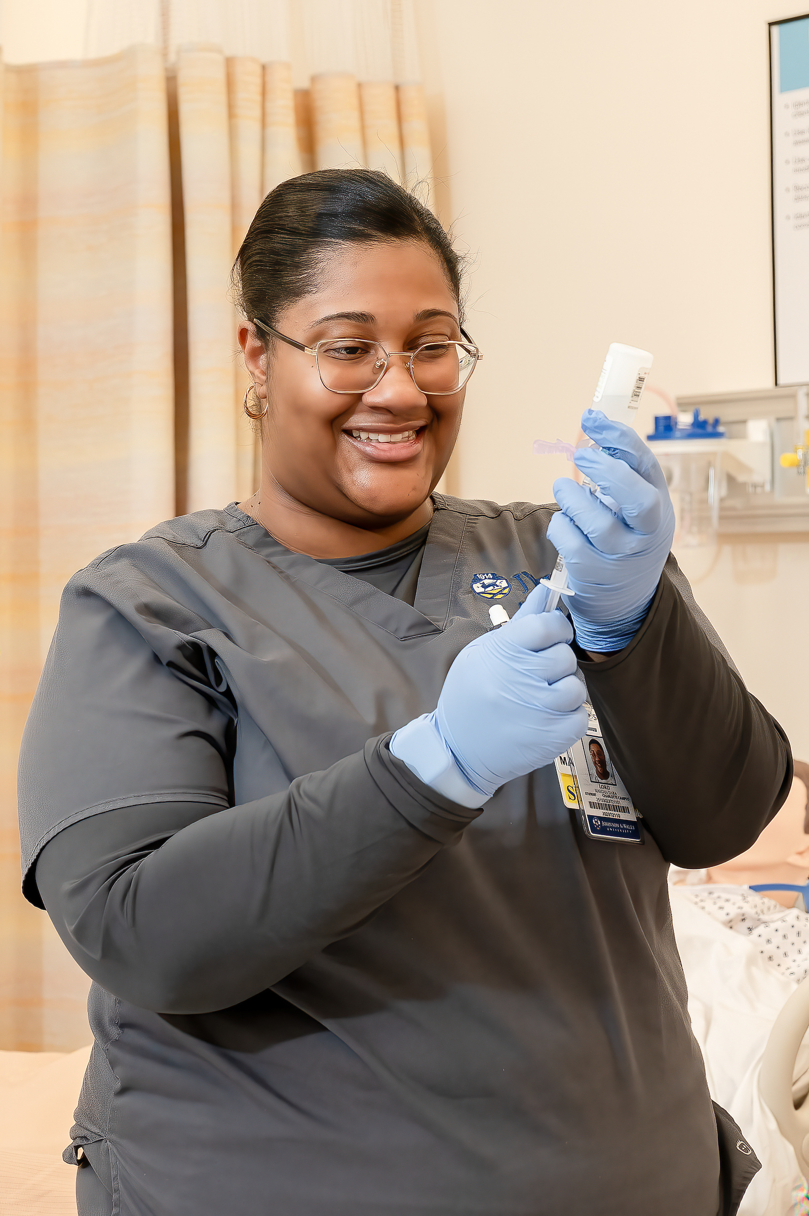 A healthcare worker in scrubs and gloves smiles while holding a syringe and vial in a clinical setting.