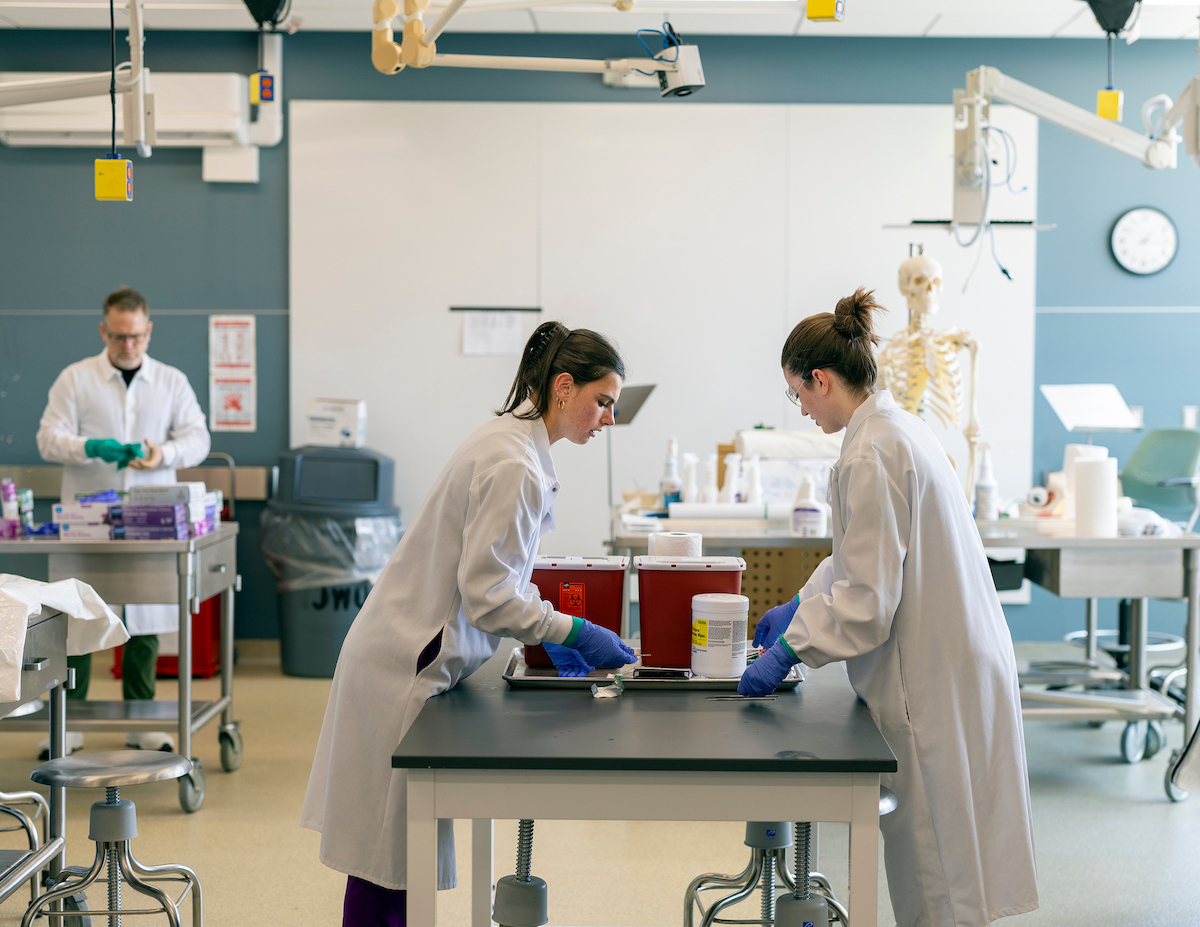 Two people in lab coats and blue gloves work together at a lab table, handling red biohazard containers and supplies in a clinical classroom setting, while another person prepares materials in the background near a skeleton model.